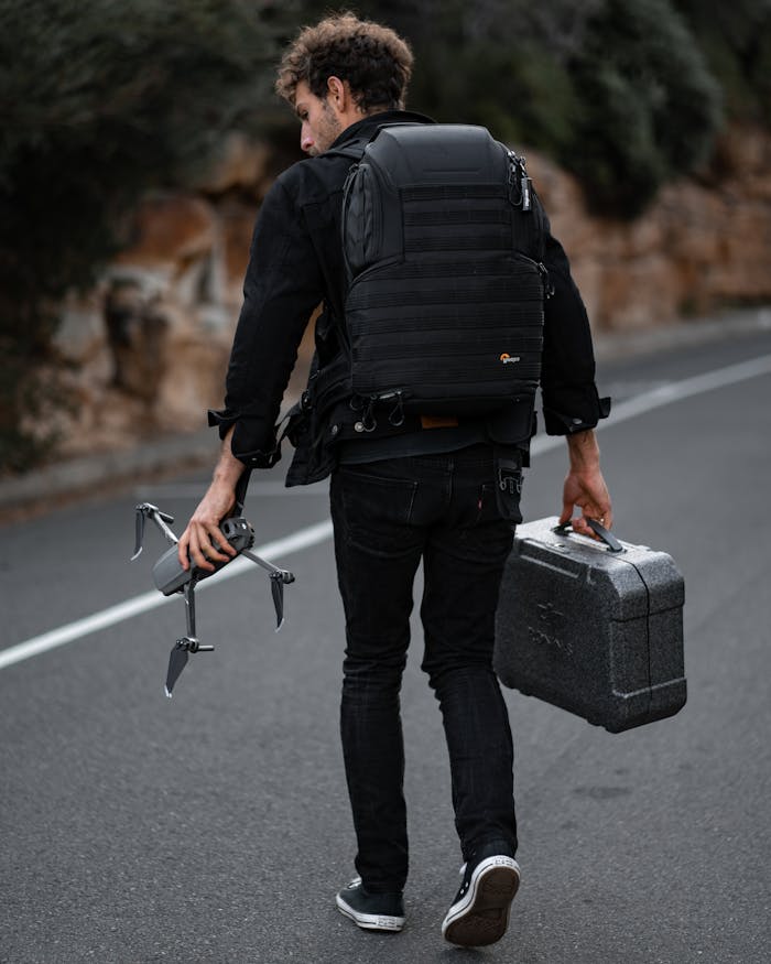 zipwp-image-6019104 A Man Walking on the Road Carrying a Gray Drone and a Case